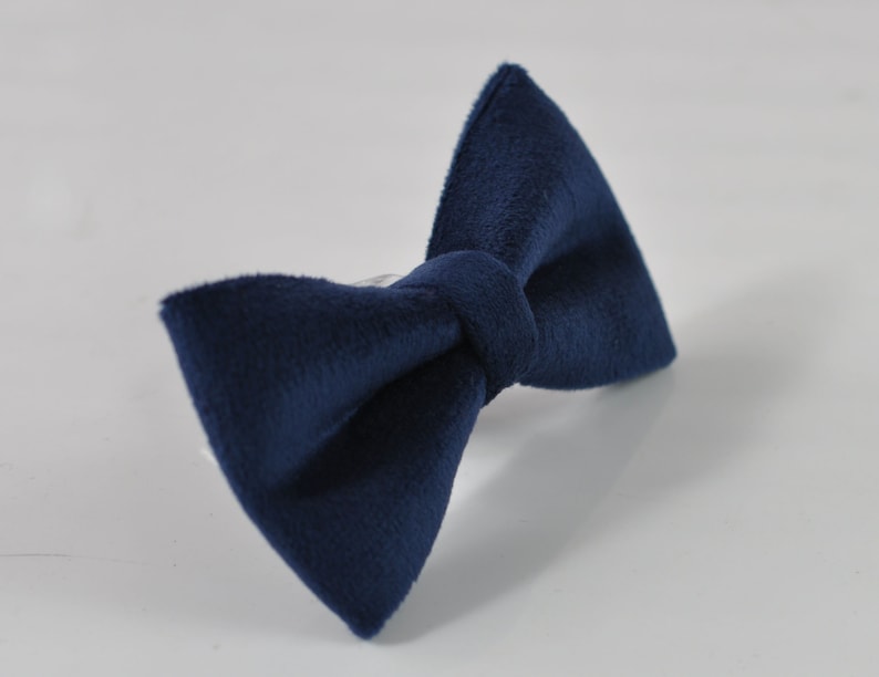 May include: A navy blue bow tie made of a soft, textured fabric. The bow tie is knotted in the center and has a classic butterfly shape. The accessory is set against a white background.