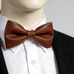 May include: A brown bow tie with a textured, leather-like finish worn over a crisp white collared shirt and a black blazer. The bow tie features a classic pre-tied shape with a central knot detail, showcasing a rich, deep brown color and grain.