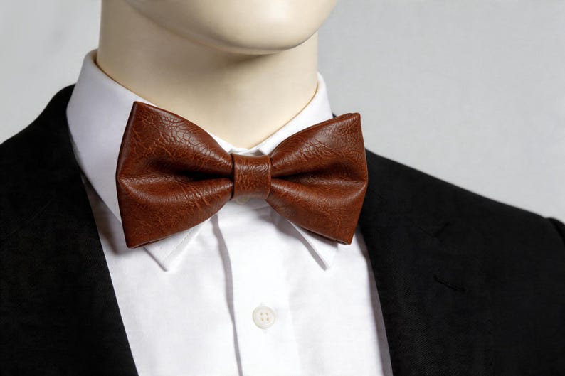 May include: A brown bow tie with a textured, leather-like finish worn over a crisp white collared shirt and a black blazer. The bow tie features a classic pre-tied shape with a central knot detail, showcasing a rich, deep brown color and grain.