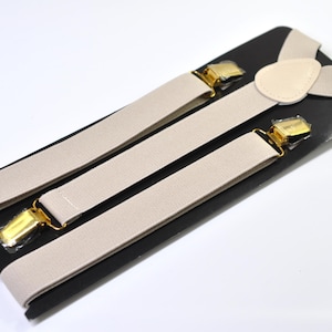 May include: Beige suspenders with gold-coloured metal clasps and a leather patch. The suspenders are displayed on a black card against a white background. A classic accessory for formal or casual wear.