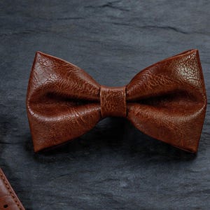 May include: A brown bow tie with a textured, pebbled surface finish. The bow tie is positioned next to a wristwatch with a matching brown leather strap and a white face, along with a pair of silver-toned metal cufflinks on the side.