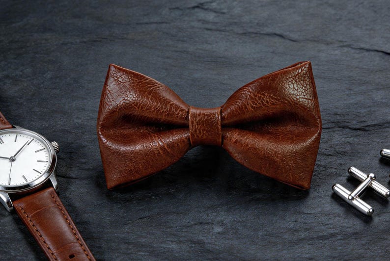 May include: A brown bow tie with a textured, pebbled surface finish. The bow tie is positioned next to a wristwatch with a matching brown leather strap and a white face, along with a pair of silver-toned metal cufflinks on the side.