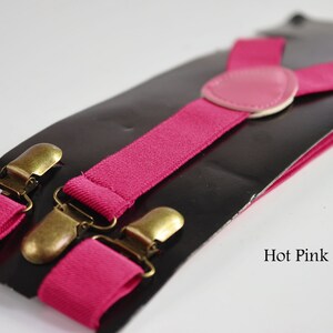 Peut inclure: Bretelles rose vif avec des clips dor&eacute;s et un patch en cuir rose. Les bretelles sont emball&eacute;es sur une carte noire avec le texte "Hot Pink".