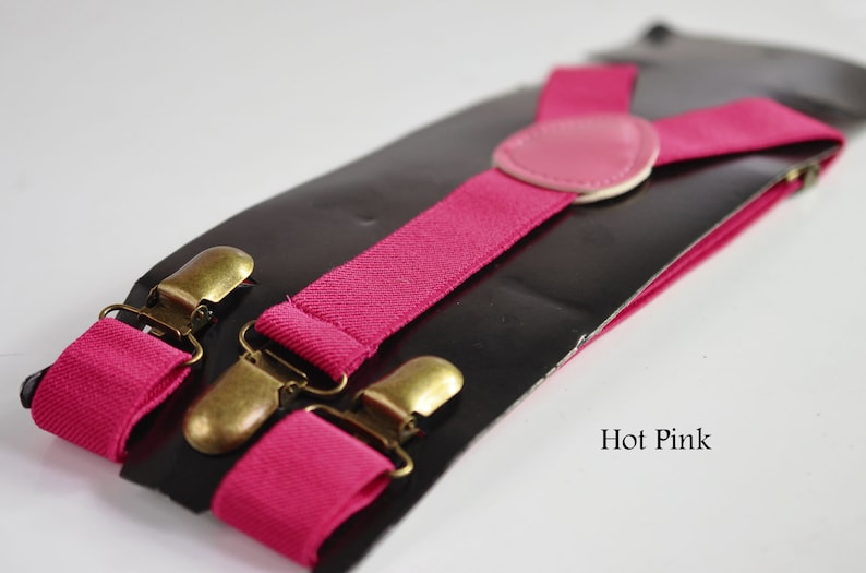 Peut inclure: Bretelles rose vif avec des clips dor&eacute;s et un patch en cuir rose. Les bretelles sont emball&eacute;es sur une carte noire avec le texte "Hot Pink".