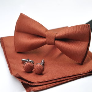 May include: A set of brown bow tie, pocket square, and cufflinks. The bow tie is made of a textured fabric and has a classic shape. The pocket square is a small square of fabric that can be folded and placed in a jacket pocket. The cufflinks are round and have a silver finish.