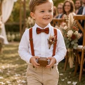 May include: A child wearing a white long-sleeved button-down shirt, tan trousers, and rust-colored suspenders. The outfit includes a matching rust-colored bow tie with teal trim, a small dried floral boutonniere, and a small wooden ring box.