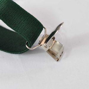 May include: A dark forest green elastic suspender strap featuring a silver-toned metal clip attachment. The clip is constructed with a hinged mechanism and a rectangular base, securely fastened to the fabric strap through a small metal loop.