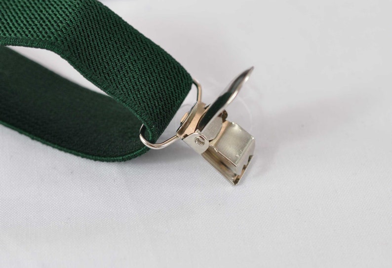 May include: A dark forest green elastic suspender strap featuring a silver-toned metal clip attachment. The clip is constructed with a hinged mechanism and a rectangular base, securely fastened to the fabric strap through a small metal loop.
