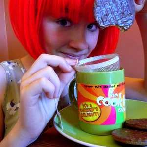 Può includere: Una donna con i capelli rosso vivo tiene in mano una tazza verde con le parole "The Cook-It Cookie Cafe" e "It's a Dunker's Delight" stampate su di essa. Sta tenendo un sacchetto di tè nella mano e sta per metterlo nella tazza. Ci sono due biscotti al cioccolato su un piatto verde davanti a lei.