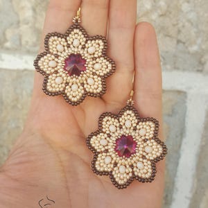 Puede incluir: Un par de pendientes de flores de cuentas con un centro de cristal rosa. Los pendientes est&aacute;n hechos con cuentas blancas y marrones y tienen un gancho dorado.