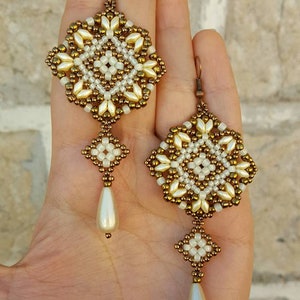 Pu&ograve; includere: Un paio di orecchini con perline dorate e bianche con un pendente a goccia di perla. Gli orecchini sono realizzati con piccole perline e presentano un design geometrico.