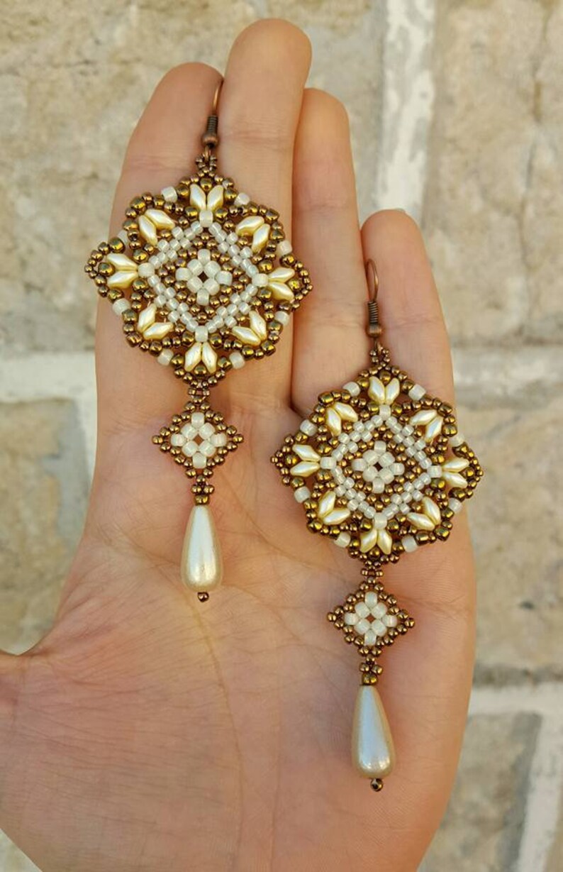 Pu&ograve; includere: Un paio di orecchini con perline dorate e bianche con un pendente a goccia di perla. Gli orecchini sono realizzati con piccole perline e presentano un design geometrico.