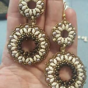 May include: A pair of gold tone earrings with two circular beaded designs. The larger circle is made of white, brown, and gold beads. The smaller circle is made of white and gold beads.