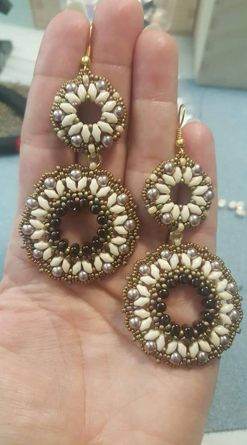 May include: A pair of gold tone earrings with two circular beaded designs. The larger circle is made of white, brown, and gold beads. The smaller circle is made of white and gold beads.