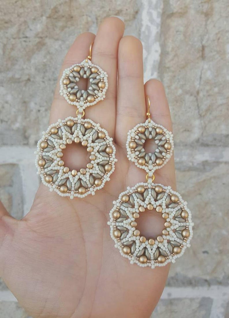 May include: A pair of gold and white beaded earrings with a circular design. The earrings are made of small beads that are arranged in a geometric pattern. The earrings are dangling and have a gold hook.