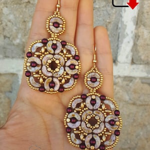 May include: A pair of gold and burgundy beaded earrings with a circular design. The earrings are made with small, round beads and have a delicate, intricate pattern.