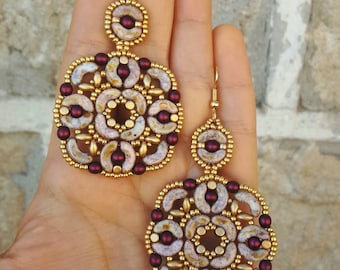 May include: A pair of gold and burgundy beaded earrings with a circular design. The earrings are made with small, round beads and have a delicate, intricate pattern.