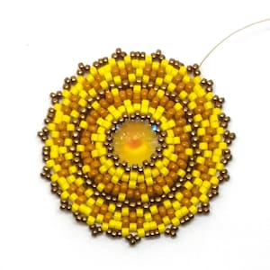 May include: A beaded sunburst design with a yellow and brown colour scheme. The centre of the sunburst features a round iridescent bead.