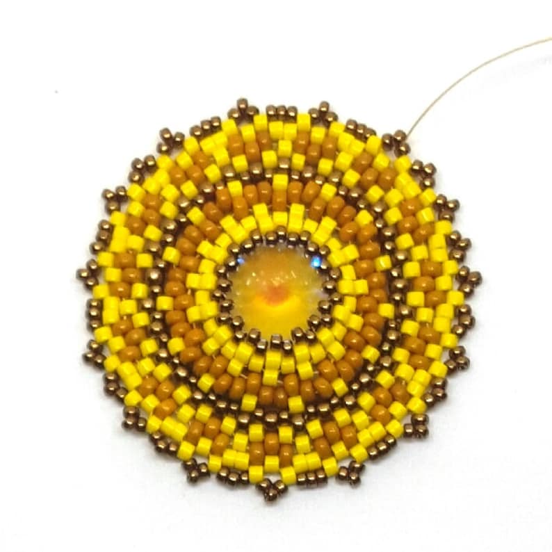 May include: A beaded sunburst design with a yellow and brown colour scheme. The centre of the sunburst features a round iridescent bead.