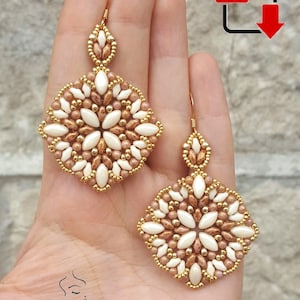 May include: A pair of gold and white beaded earrings with a square shape and a floral design. The earrings are hanging from gold hooks.