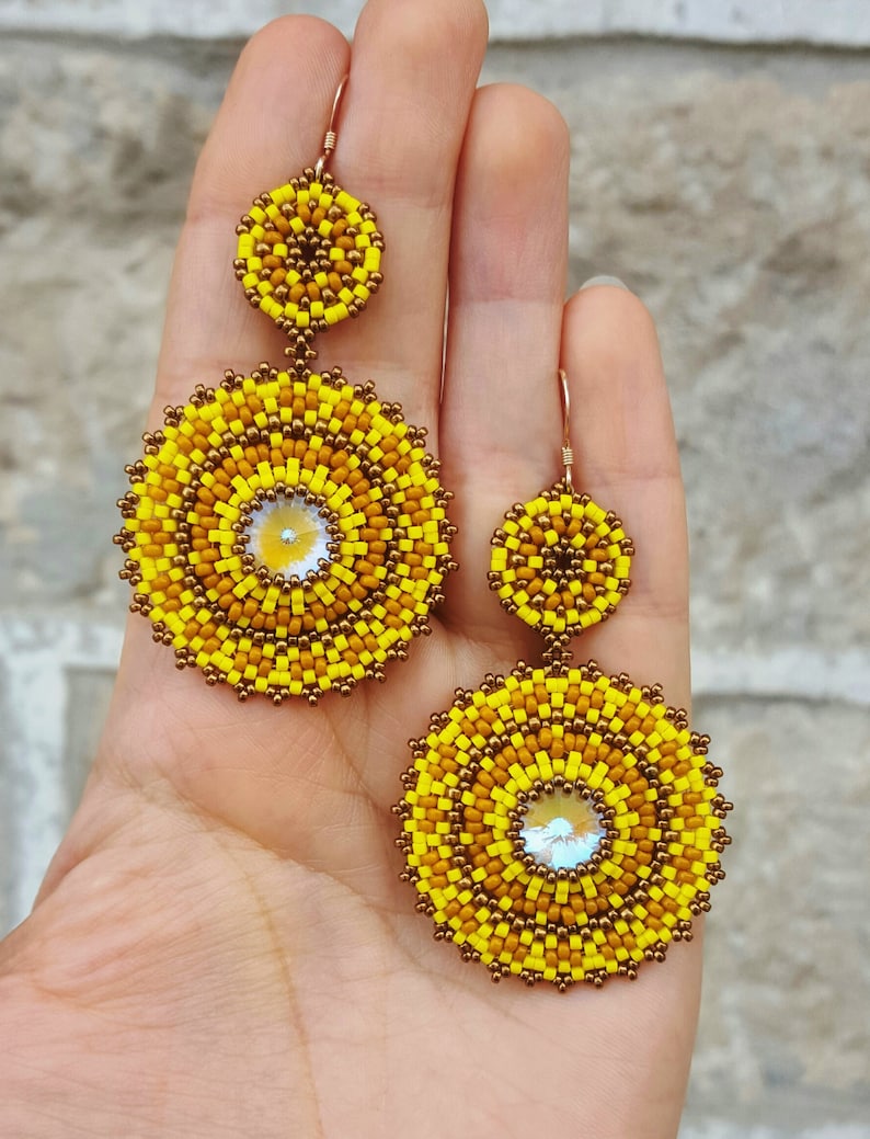 May include: A pair of yellow and brown beaded earrings with a round, layered design. Each earring has a smaller round bead at the top and a larger round bead at the bottom. The beads are made of small, colourful beads.