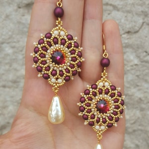 Peut inclure: Une paire de boucles d'oreilles en perles avec un motif de couleur bordeaux et or. Chaque boucle d'oreille pr&eacute;sente un motif rond en perles avec un cristal rouge au centre et une goutte nacr&eacute;e suspendue en dessous.