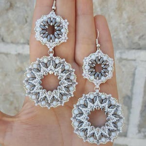May include: A pair of silver dangle earrings with two beaded circles, one larger than the other. The circles are made of white and gray beads.