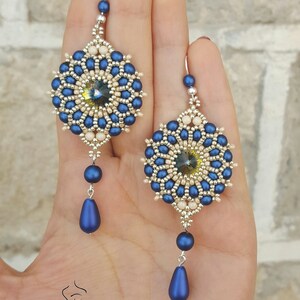 Peut inclure: Une paire de boucles d'oreilles en perles bleues et cr&egrave;me avec un pendentif en forme de larme. Les boucles d'oreilles pr&eacute;sentent un motif rond en perles avec un centre en cristal.