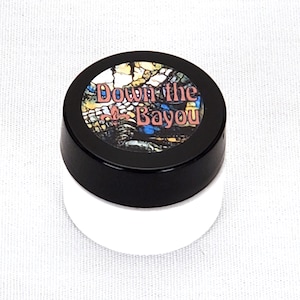May include: A small white jar with a black lid. The lid has a colorful sticker with the text "Down the Bayou" and a fleur-de-lis.