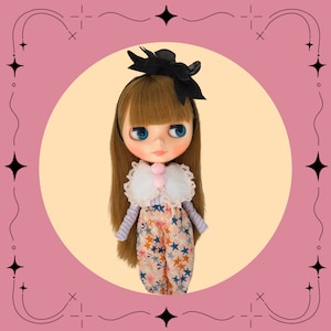 Könnte beinhalten: Eine Puppe mit langen braunen Haaren, blauen Augen und einem rosa-weißen Rüschenkragen. Sie trägt ein rosa-blaues gemustertes Kleid mit Sternen und ein schwarzes Stirnband mit Schleife.