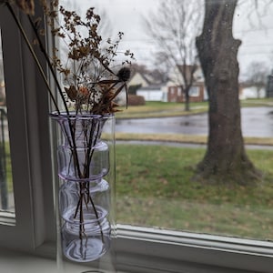 May include: A clear glass vase with a layered design in shades of purple, filled with dried flowers and branches. The vase is sitting on a windowsill with a view of a tree and houses in the background.