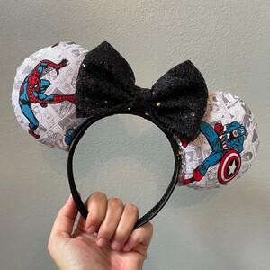 May include: A pair of black and white fabric mouse ears with a black bow. The ears feature comic book images of Spider-Man and Captain America.