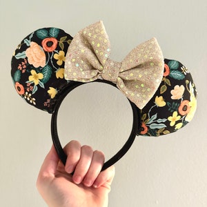 May include: Floral print Minnie Mouse ears headband with a gold glitter bow. The ears are black with a floral pattern in shades of pink, yellow, green, and orange.