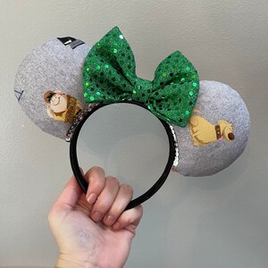 Puede incluir: Orejas de ratón de tela gris y blanca con un lazo de lentejuelas verde. Las orejas muestran un perro y un personaje de la película Up.