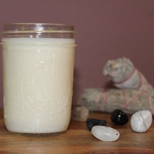 Op de afbeelding: Een witte kaars in een glazen pot met een wit label. De kaars staat op een houten oppervlak met een bundel salie, een zwarte steen en een witte steen.
