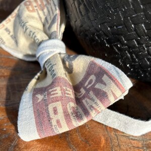 May include: A bow tie made from fabric with a newspaper print. The bow tie is tied and has a white band around the center. The fabric has text that reads "Bourbon" and "Rye".