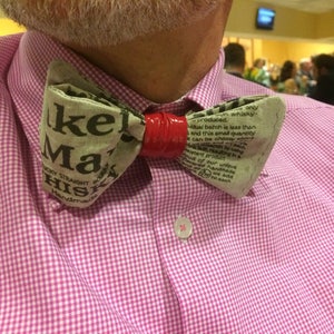 May include: A bow tie made from a piece of fabric with the words "Make Mai His" printed on it. The bow tie is red and white and is tied around a person's neck.
