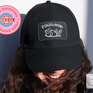 May include: Black baseball cap with a rectangular patch displaying the word "POGUELANDIA" and a quirky white illustration. The cap is made of a sturdy material and has a curved peak. A fun accessory for casual wear.