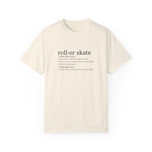 May include: A beige t-shirt with a black graphic design that defines the word "roller skate" as a type of boot with four wheels and a verb meaning to glide across a hard surface on roller skates.