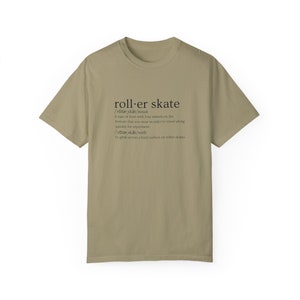 May include: A khaki t-shirt with a black graphic print of the word "roller skate" defined as a type of boot with four wheels on the bottom that you wear to travel quickly for enjoyment. The definition also includes the verb "roller skate" defined as to glide across a hard surface on roller skates.