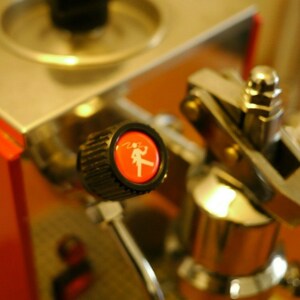 May include: Close-up of a silver espresso machine with a red and black knob. The knob has a white silhouette of a person holding a cup.