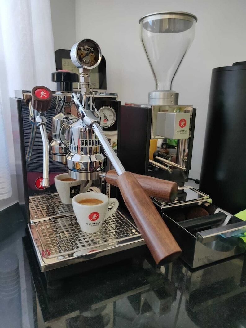 May include: A silver espresso machine with two cups of espresso on a drip tray. The machine has a wooden handle and a pressure gauge. The machine is labeled "OLYMPIA EXPRESS".