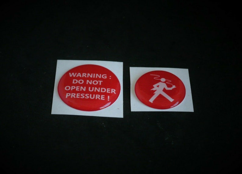 May include: Two red round stickers with white text and a white graphic. The first sticker says "WARNING: DO NOT OPEN UNDER PRESSURE!" The second sticker has a white graphic of a person walking with a tray.