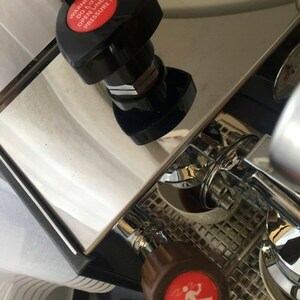 May include: Close-up of a silver espresso machine with a red button that reads "WARNING DO NOT OPEN UNDER PRESSURE!" and a brown knob with a red warning symbol.