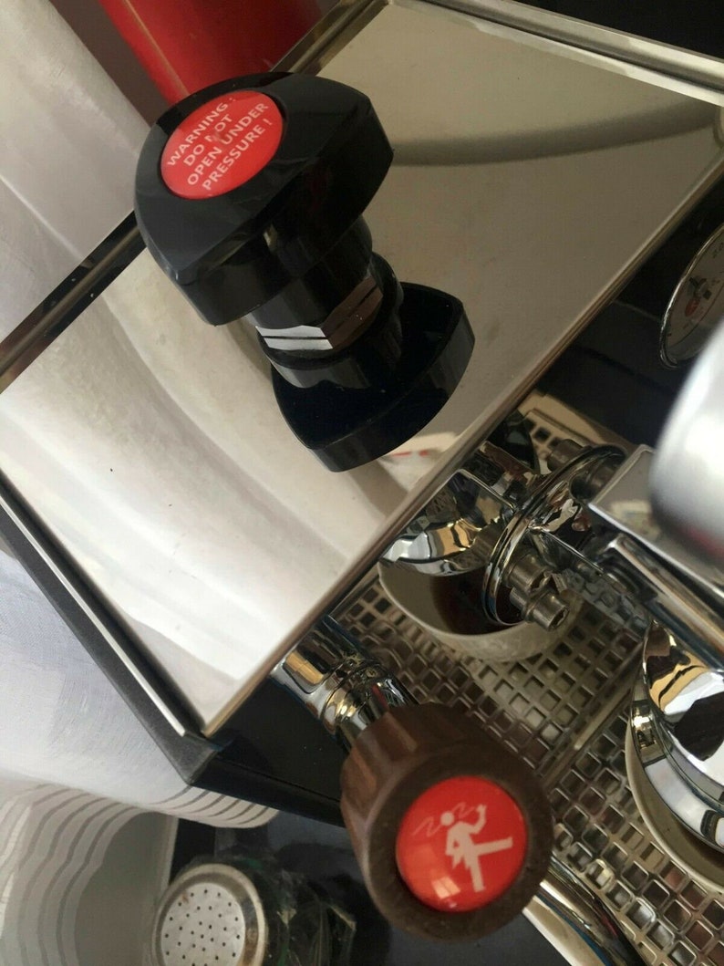 May include: Close-up of a silver espresso machine with a red button that reads "WARNING DO NOT OPEN UNDER PRESSURE!" and a brown knob with a red warning symbol.