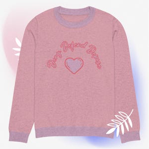 Op de afbeelding: Roze longsleeve trui met een roze hart en de tekst "Deny Defend Deplore" in roze schrift.