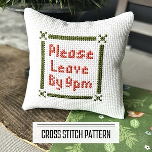 A DIY cross stitch pattern for a mini pillow that can be used as a pin cushion. The finished pillow is sitting on a wooden board.