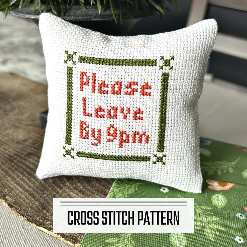 A DIY cross stitch pattern for a mini pillow that can be used as a pin cushion. The finished pillow is sitting on a wooden board.