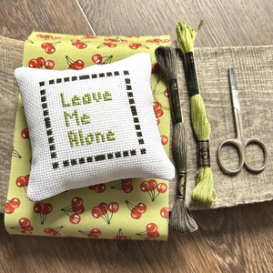 The finished pillow, made from this cross stitch pattern, is sitting on a board with all of the supplies.