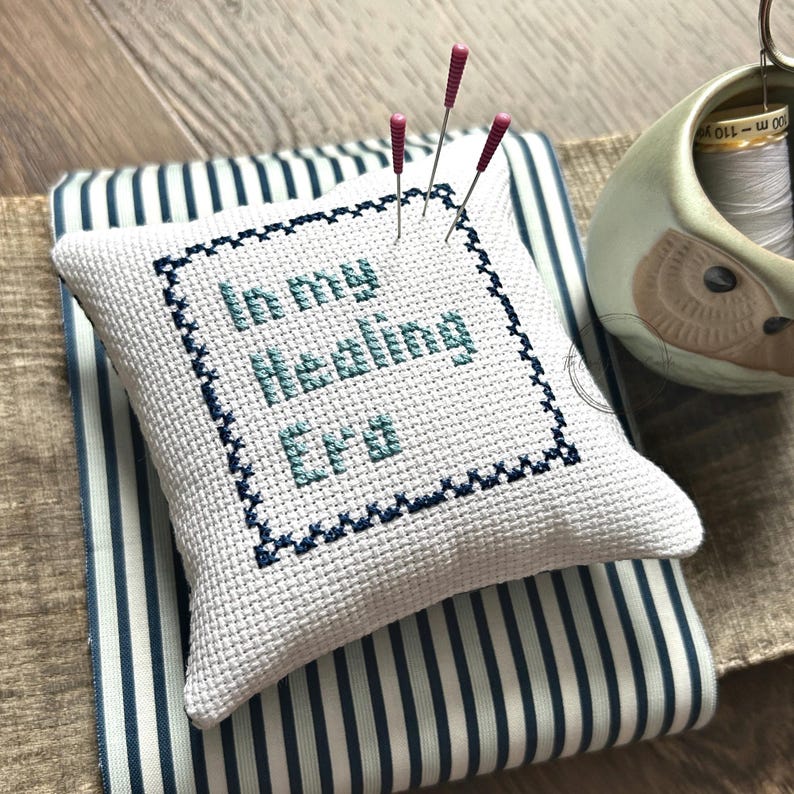 The finished pillow, made from this cross stitch pattern, is sitting on a wooden board with pins sticking out.